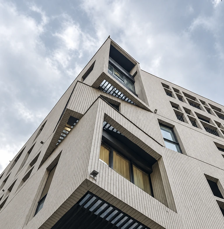 Upward view of Konj Residence Farmaniye with pale vertical tiles, projecting angular frames and deeply recessed windows beneath a cloudy sky
